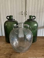 Three vintage glass bottles shown together: two green glass bottles with loop handles near neck and one clear glass bottle with textured horizontal segments and double loop handles