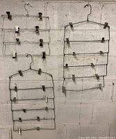 Lot of 3 vintage metal wire hangers with multiple tiers and clips hanging on a wall for display.