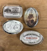 Four glass paperweights on wooden surface, showing various vintage advertising and artwork