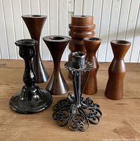 Group photo showing seven candle holders with different styles and materials, placed on wooden surface against white wall.