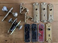 Top-down view of vintage door hardware including doorknobs, backplates in brass, black, red, and green finishes, and decorative brackets.
