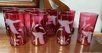Set of eight cranberry glasses with etched bird and reed motif shown in a group on a wooden surface.