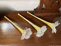 Three vintage ART LASI glass flowers displayed side by side on wooden surface showing full flower with long yellow stems and clear ruffled bell-shaped blossoms
