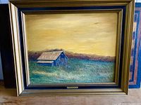 Framed oil painting showing a rustic barn with prairie grass and trees in the background, set under a yellow sky.