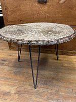 Side view of the vintage side table showing the woven wicker top and three hairpin metal legs on a wood floor.