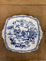 Full view of the square blue willow plate showing detailed oriental scene with pagodas, birds, bridge, and boat in blue on white with floral border and small handles on sides.