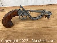 Photo showing a pewter flintlock pistol replica with intricate metalwork and a wooden handle placed on a wooden surface.