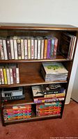 Wooden shelf unit filled with audio cassettes, CDs stacked on one shelf, portable cassette players, and VHS tapes on bottom shelf. Visible scratches on shelf surface.