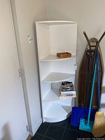White laminate corner shelf with four shelves, showing triangular shelves and placement in corner near wall and household items beside it.