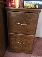 Wooden 2-drawer file cabinet with brass handles. Bottom drawer broken as reported. Cabinet wood grain visible with some signs of wear.