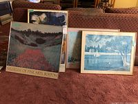Four framed pieces of wall art leaning against furniture showing two Museum of Fine Arts posters in front, one with a landscape in red and green tones, one with text 'MUSEUM OF FINE ARTS, BOSTON'.