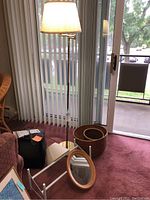 Photo showing the adjustable floor lamp with pleated beige shade, oval wood-framed mirror leaning against the lamp base, small white metal rack, second lamp shade on the floor, and two round woven baskets on a pink carpet near sliding glass doors.
