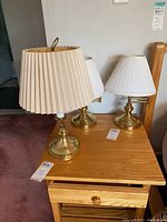 Photo showing three brass tone lamps with pleated fabric shades on a wooden nightstand, two lamps 15 inches tall with swing arms visible.