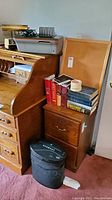 Photo showing office items on a wooden roll-top desk and a small filing cabinet with books, lamp, printer, shredder and office supplies