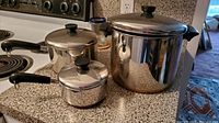 Three stainless steel pots on kitchen counter showing small and medium saucepans with lids and large stock pot with lid and strainer insert