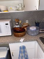 Two wooden salad bowls, a salad spinner, ceramic pot, and cooking utensils in a white pitcher on a grey kitchen countertop next to a microwave