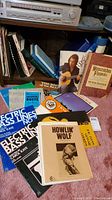 Photo of multiple music and instructional books stacked and leaning, showing titles like 'Henscratches Flyspecks' by Peter Seeger, Howlin' Wolf, and 'Electric Bass Lines' by Carol Kaye.