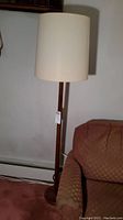 Full view of vintage wooden floor lamp standing next to upholstered chair, showing lamp and shade.