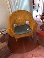 Front angle view of the wicker rocking chair with blue cushion on a reddish carpet among other upholstered furniture.