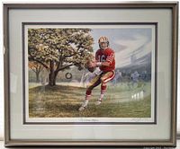 Full view of framed lithograph showing Joe Montana in mid-action on a football field, with natural scenery and background figures, enclosed in a dark wood frame.