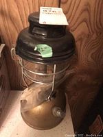 Full view of the lantern showing the brass base, glass globe, black top, and metal wire cage with instruction tag attached.