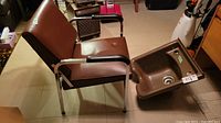 Side view of vintage brown vinyl and chrome hairdresser chair showing folding seat and black armrests, next to the brown plastic sink.