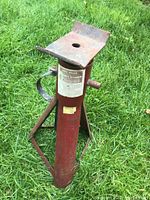 Photo showing the red automotive jack stand standing on grass with visible rust and wear, triangular base and adjustment handles