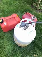 Two red plastic jerry cans one larger with cap and one smaller missing cap, and an empty white propane BBQ tank with attached hose and valve, all pictured outdoors on green grass.