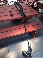 Side view of black Jobmate electric weed wacker on wooden picnic bench, showing long shaft and curved cutting head with protective shield.