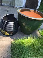 Overall view of green glazed terracotta plant pot next to coiled black plastic garden edging on concrete and grass background.