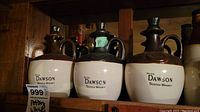 Set of three pottery jugs on shelf, two-tone brown and cream glazing, labeled Peter Dawson Scotch Whisky