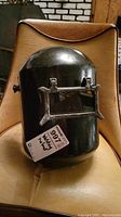 Front view of black plastic Sure Guard welding helmet placed on a brown leather chair, showing surface scratches and flip-up viewing window with darkened lens.