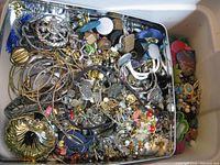 Metal tin filled with assorted costume jewelry, including bangles, earrings, pins, chains, colorful plastic and enamel parts, showing an unused pile of mixed condition items.