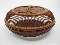 Front angled view showing the round shape, open weave design, and lidded top with central handle wrapped in golden cord.