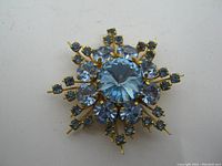 Close-up front view of blue snowflake brooch pin showing large central faceted blue gemstone and surrounding smaller stones set in gold-tone metal.
