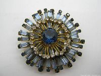 Front view showing large central blue faceted rhinestone, surrounded by clear rhinestones and outer layer of light blue baguette and small round blue rhinestones set in gold-tone metal.