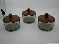 Three glass condiment dishes with teak wood lids shown together, front angle