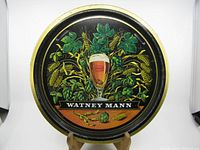 Full view of the round metal Watney Mann beer tray showing colorful hop and barley design, glass of beer, and branding.