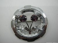 Front view of the Mizpah Scottish brooch featuring round circular frame with two purple stones and silver-tone thistle leaves design.