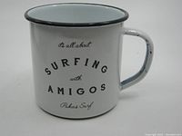 Front view of white enamel tin cup with black rim and Pukas Surf surfing related text.