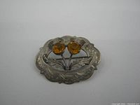 Front view of a silver oval brooch with scalloped edges, amber-colored stones, and leaf engravings.