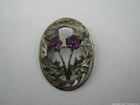 Front view of an oval sterling silver brooch featuring two Scottish thistles with purple faceted stones as flowers.
