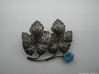 Front view of the sterling silver leaf brooch showing three detailed leaves and blue faceted round stone attached to one stem.