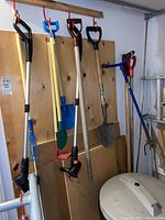 Full view of hanging garden tools including two weedeaters, hoe, shovel, rake, and wooden handles.