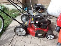 Overall side view of the red Yard Machines push lawn mower showing its body, wheels, and handle, placed outdoors on a paved surface near a wall.