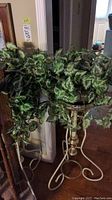 Photo showing two metal plant stands with faux ivy plants. One stand is white with gold accents, the other gold. Both have curved scrollwork bases and hold bushy faux ivy plants.
