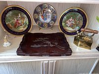Photo showing three decorative Limoges Fragonard plates, one wood engraved tray, and a vintage metal iron doorstop with wooden handle arranged on shelf