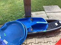 View showing one black Ho2! racer toboggan and two blue circular saucer toboggans stacked together outdoors on stone paving.