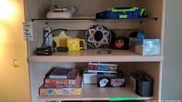 Wide photo showing several shelves holding multiple board games, electronic game devices, hats, decorative stones, a Mario-themed mug, and a Nexttech microphone.