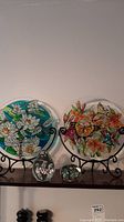 Two circular art glass plates with painted nature scenes displayed on black metal easels with two small art glass paperweights in front.